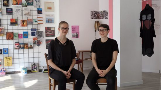 Anna Erdmann und Franziska Goralski vor dem großen Bücher- und Magazinregal und Textilarbeiten von Nada van Dalen aus Rotterdam