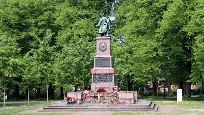 Mahnmal mit künstlerisch gestalteter Spange, ohne Fragilitätsschild, jedoch mit Blumen.